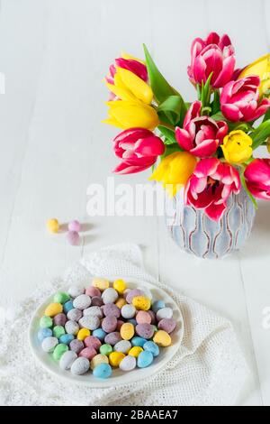 Festive Easter candies covered eggs in various pastel colors on the plate with vintage napkin near vase with fresh tulips on wooden table. Family holi Stock Photo