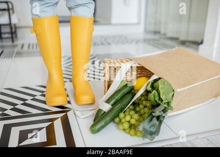 Shopping bag full of fresh vegetables and greens which fell on the floor in the corridor at home, woman in yellow rubber boots Stock Photo