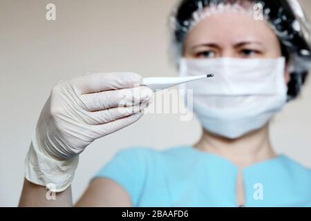 Coronavirus symptoms, woman in medical mask measures body temperature. Doctor looks at digital thermometer in her hand, concept of cold and flu Stock Photo