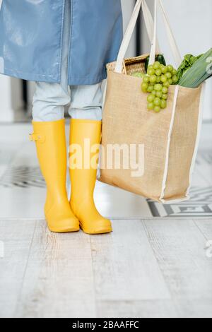 Woman in yellow rubber boots and raincoat standing with shopping bag full of fresh food in the corridor. Woman coming home in rainy weather Stock Photo
