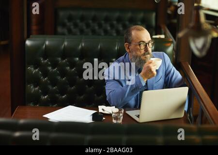 Senior bearded businessman drinking coffee and thinking about something while working with laptop in cafe Stock Photo