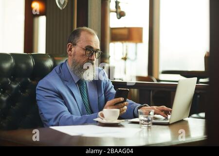 Senior businessman with beard and in eyeglasses sitting at the table drinking coffee and using laptop in cafe Stock Photo