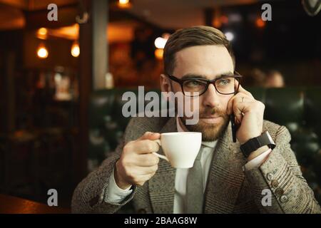 Young bearded businessman in eyeglasses talking on mobile phone and drinking coffee in coffee shop Stock Photo