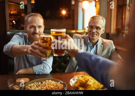 Young bearded men holding glasses of beer and toasting with them while having fun in the bar Stock Photo