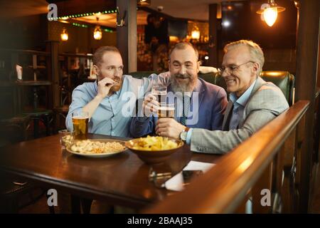 Three mature men sitting at the table drinking beer with snacks and talking in the pub Stock Photo