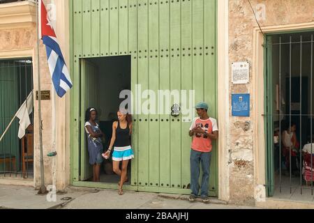 Cuban man standing outside office and woman inside office both looking at young woman standing in doorway in Havana Stock Photo