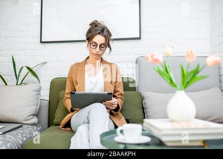Young creative and well-dressed woman working on a digital tablet while sitting on the comfortable couch at home. Concept of a freelance and creative work online Stock Photo
