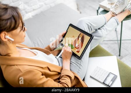 Young creative woman working as a web designer or artist, drawing on a digital tablet while sitting on the comfortable sofa at home Stock Photo