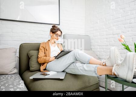 Young creative and well-dressed woman working on a digital tablet while sitting on the comfortable couch at home. Concept of a freelance and creative work online Stock Photo