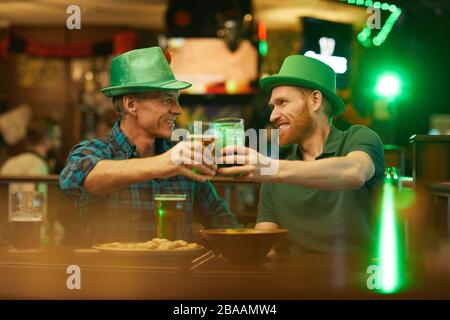 Two friends in green hats toasting with glasses of beer while have the meeting in the pub Stock Photo