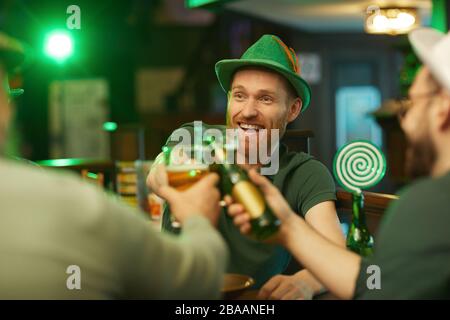 Happy bearded man in green hat having fun with his friends in the bar they toasting with glasses of beer Stock Photo