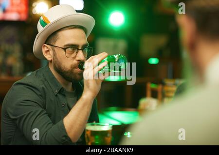 Young bearded man in eyeglasses and in hat drinking beer from the bottle while sitting in the pub Stock Photo