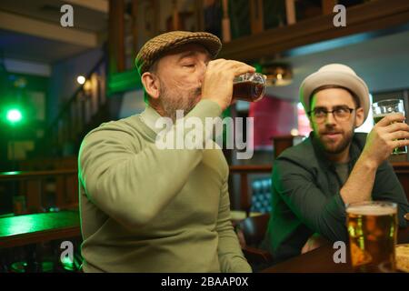 Mature bearded man tasting beer while resting in the bar together with his friends Stock Photo