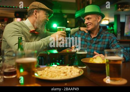 Two smiling friends talking and toasting with beer while sitting at the table with snacks in the bar Stock Photo