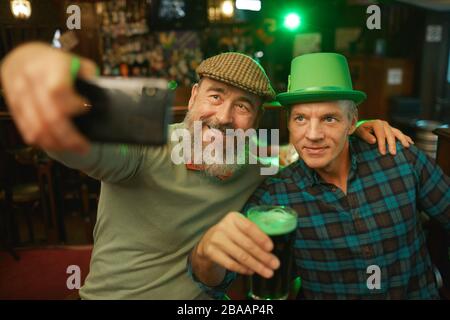 Two mature men in hats taking selfie portrait on mobile phone while drinking beer in the pub Stock Photo