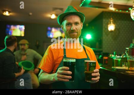Portrait of young bearded waiter in green apron and hat holding two glasses of beer and smiling at camera while working in the pub Stock Photo