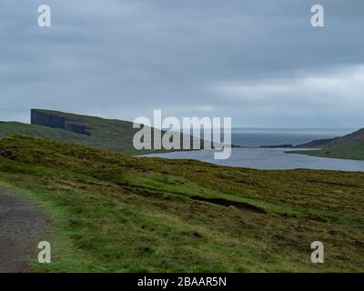 Faroe Islands. Trælanípan or Slave cliffs on Vagar Island. Green grass fields and sheeps in the foreground. Lake Leitisvatn in the background. Hazy la Stock Photo