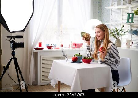 Food blogger cooking fresh vegan salad of fruits in kitchen studio, filming tutorial on camera for video channel. Female influencer holds apple Stock Photo
