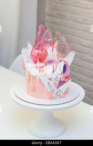 Modern elegant pink cake with caramel, white chocolate, waffle paper, meringues and french macaroons decoration on white cakestand. Stock Photo