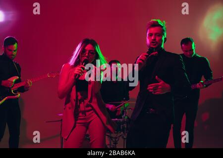 Young and joyful caucasian musicians performing on red studio background in neon light. Concept of music, hobby, festival. Colorful portrait of modern artist. Attented and inspired. Art, cover band. Stock Photo