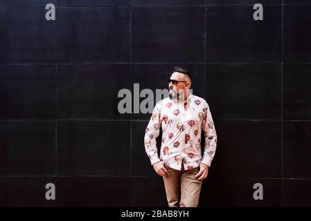 Bearded man wearing sunglasses leaning on black wall, looking aside Stock Photo