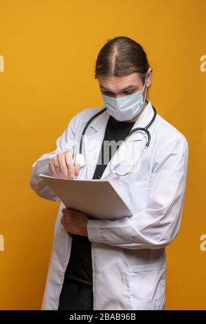 Young doctor taking notes on his map while wearing mask protection against Covid-19 virus. Stethoscope displayed around his neck Stock Photo