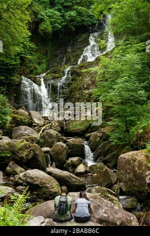 Torc Waterfall in Killarney young couple of tourists sitting on boulder and enjoying the view at the Killarney National Park, County Kerry, Ireland Stock Photo