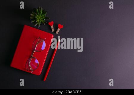 The red notebook stands on a black background, on the notebook there are glasses and next to the notebook, a flower, two clothespins in the shape of Stock Photo