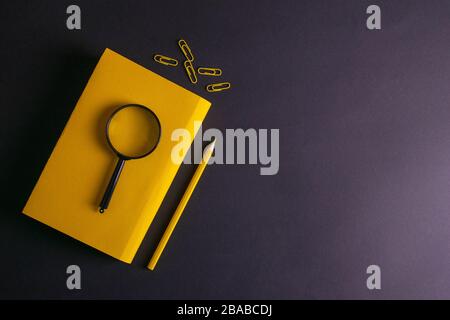 Yellow pencil, yellow notebook glasses, pins, clip paper and a magnifying glass on on black background Stock Photo