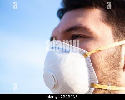 Man with mask to protect her from Corona virus looking to the side hopeful blue sky on the background. Handsome brunette dark haired guy with medical Stock Photo
