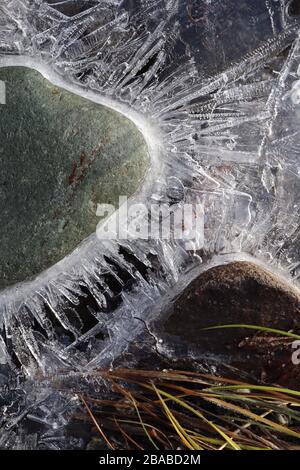 Closeup of semi-transparent rays of ice crystals around submerged rock with a fall leaf, on a small creek early morning. Stock Photo