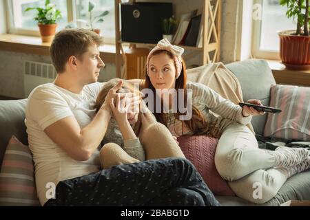 It's not for kids. Family spending nice time together at home. Looks wondered. Mom, dad and daughter having fun, eating pizza, watching TV. Togetherness, home comfort, love, relations concept. Stock Photo