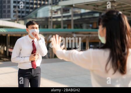 Asian ill businessman cough with mask and businesswoman stop sign hand him to keep distance protect from COVID-19 viruses and people social distancing Stock Photo