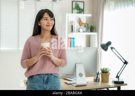 Asian beautiful young woman work from home working on computer and drinking coffeewhile relax from her work standing in living room at home. Social di Stock Photo