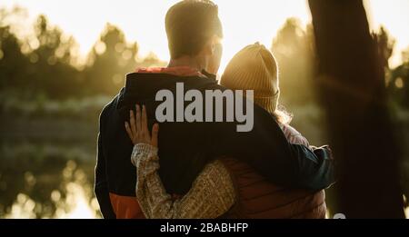 Rear view of couple standing together by the lake. Man and woman in warm clothing looking at a view. Stock Photo