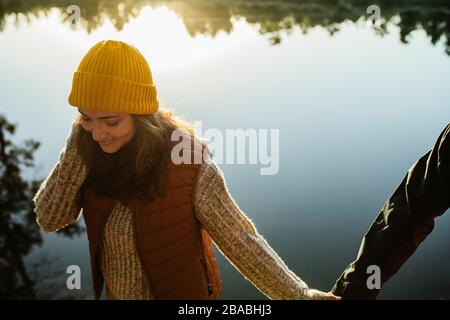 Woman in warm clothing walking holding hand of her man. Couple walking by the lake. Stock Photo