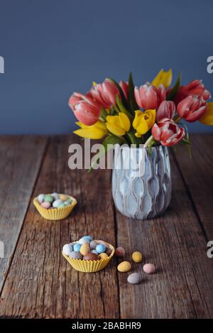 Easter candies covered eggs in various pastel colors in paper baskets near vase with fresh tulips on rustic wooden table and dark blue background. Fas Stock Photo