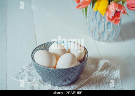 Farm Fresh white eggs in ceramic grey bowl on vintage napkin and vase with fresh pink and yellow tulips on the white wooden table. Natural Easter conc Stock Photo
