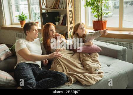 Family spending nice time together at home, looks happy and excited. Mom, dad and daughter having fun, eating pizza, watching sport championship on TV. Togetherness, home comfort, love concept. Stock Photo