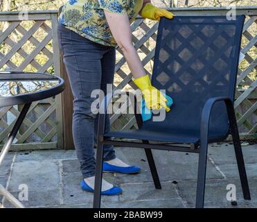 A female wipes clean a mesh garden chair with a microfibre cloth. She is wearing yellow rubber gloves. Stock Photo