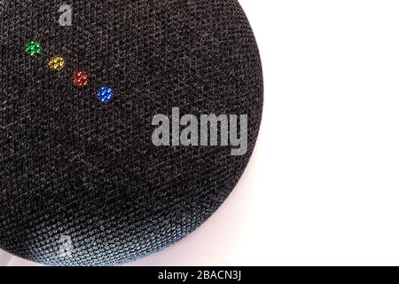 A dark grey Google Nest Mini smart speaker featuring Google Assistant sits on a white surface. Its lights show the colors of Google's brand and logo. Stock Photo