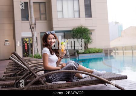 Beautiful young Asian woman holding juice bottle while relaxing in deck chair near the pool. Stock Photo