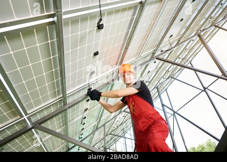 Solar power station with large volumes. Young man inside her is setting up a system, smiling to the camera. Worker in an orange uniform and a helmet on his head. Solar station development concept. Stock Photo