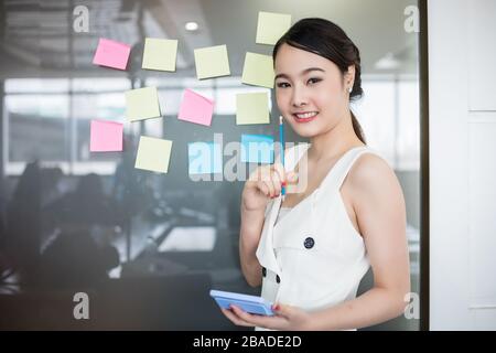 Asian business women smiling and Business people meeting at office and use post it notes to share idea. Brainstorming concept. Sticky note on glass wa Stock Photo