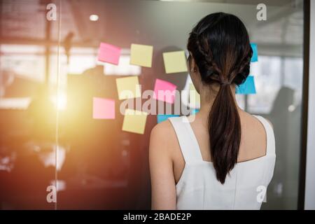 Asian business women smiling and Business people meeting at office and use post it notes to share idea. Brainstorming concept. Sticky note on glass wa Stock Photo