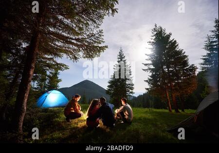 Loving couple surrounded by friends at an evening camping in the mountains. People sit by the fire near the tents under the spruce trees against the backdrop of the mountain valley. Stock Photo