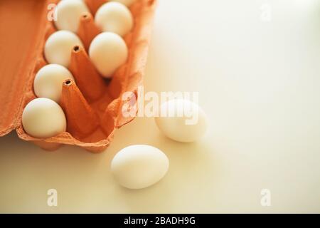 On a white light table is an orange cardboard package with white chicken eggs and two eggs are lying next to it on the table. Stock Photo