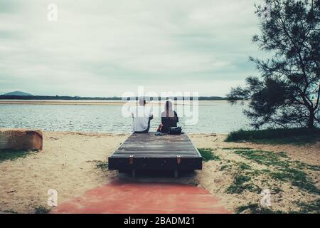 Young couple sitting together on a bench and looking at the river. Back view. Stock Photo