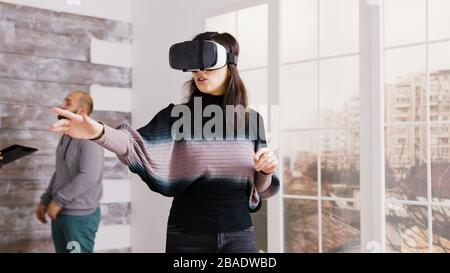 Female architect using virtual reality goggles in empty apartment and real estate agent talking with client in the background. Stock Photo