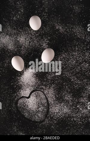 Low key image of a white eggs and a heart shaped imprint on black background sprinkled with flour. Cooking concept. Backdrop for your design. Flat lay Stock Photo